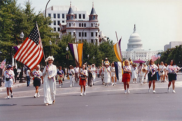 The Iowa State contingent at the 75th anniversary suffrage parade in Washington, DC, in 1995, with the U.S. Capitol in the background. Many of the women – including Dr. Betsy Hoffman, who is at the front of the group and carrying a large U.S. flag – are dressed in the white clothing that suffragists were known for. Two women carry the yellow-and-purple banners of the suffragists.