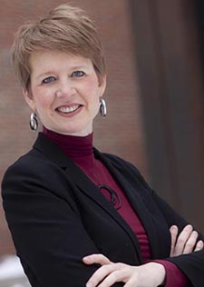 Dr. Lauren Hughes - head-and-shoulders photo of a white women with short red hair and silver hoop earrings, wearing a dark blazer over a wine-colored turtleneck, standing with her arms crossed over her chest