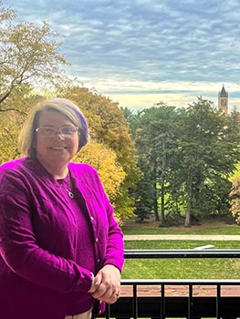 Photo of Karen Kedrowski on the balcony at Catt Hall, with a cloudy sky, trees and the top portion of the Campanile visible behind her. Kedrowski is wearing a magenta sweater set.