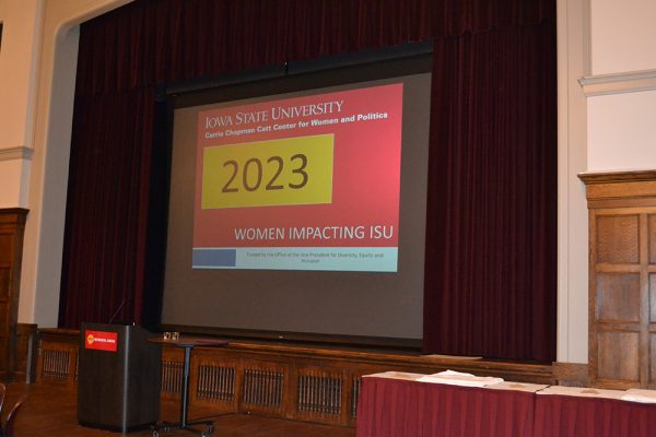 The stage area at the front of the Great Hall. A podium and two tables holding the 2023 Women Impacting ISU calendars, covered by white cloths, are in front of the stage. The screen is pulled down in front of the curtain and a slide saying "2023 Women Impacting ISU" is displayed.