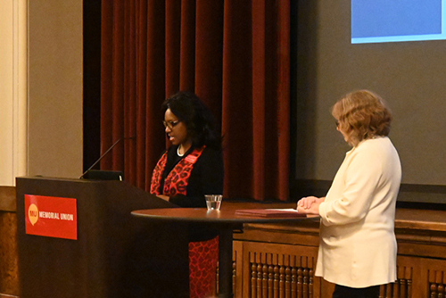 Dr. Sharon Perry Fantini, a Black women wearing a red-and-black outfit. stands at the podium in front of the stage and is greeting the reception guests, and Sue Cloud, a white woman wearing a white sweater jacket and black pants, stands nearby.