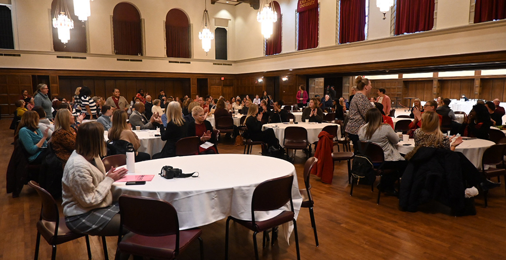 Reception guests are seated at white cloth-covered tables in the Great Hall. About a dozen women are standing while the others applaud.