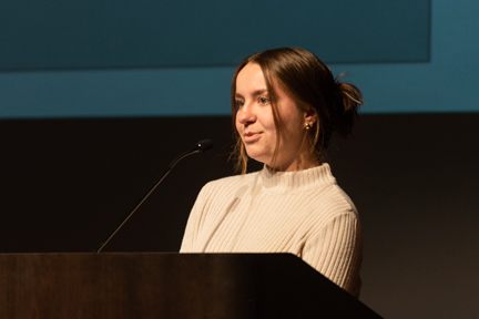 Head-and-shoulders photo of Kathryn Burns, a young woman with brown hair pulled into a bun, wearing a cream-colored mock turtleneck sweater and standing at a podium to introduce a speaker.