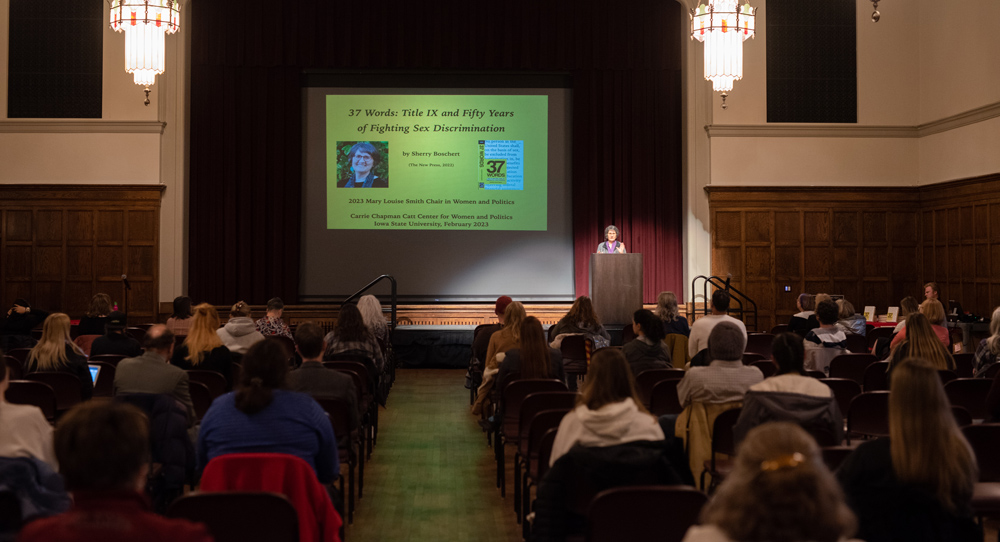 Photograph of Sherry Boschert at the podium in the Great Hall, with the first several rows of the audience visible. The first slide of her presentation is visible on the screen behind her.