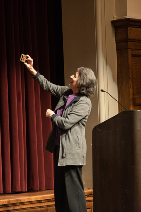 A photo of Sherry Boschert, a middle-aged woman wearing a purple blouse and grey jacket, turning to take a selfie with the audience at her presentation.