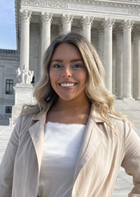 Head-and-shoulders photo of a smiling young woman with shoulder-length blonde hair, wearing a light-colored jacket over a white blouse, standing in front of the U.S. Supreme Court building.
