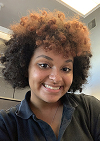 Head-and-shoulders photo of a smiling young woman with a brown and orange afro, wearing a dark blouse. Behind her you can see some of an office cubicle.