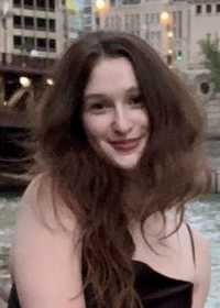 Head-and-shoulders photo of a smiling young woman with long brown hair, wearing a black spaghetti-strap dress. In the background is a city scene with a canal.