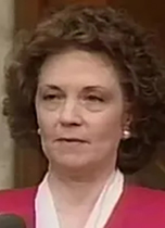 A head-and-shoulders photo of a middle-aged white woman with short curly brown hair, wearing a deep pink blazer and white blouse.