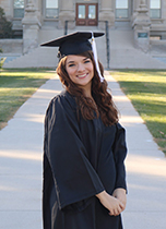 Young women with long brown hair in a black graduation robe and cap, standing on the sidewalk outside an academic building.