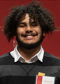 Head-and-shoulders photo of a smiling young man with a brown afro and a trim mustache and beard, wearing a two-toned gray sweater over a white shirt. The background is red.
