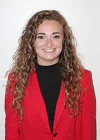Head-and-shoulders photo of a smiling young woman with long curly light brown hair, wearing a red blazer and black top. The background is plain white.