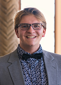 Head-and-shoulders photo of a young man with short blonde hair and glasses, wearing a grey blazer, blue-and-white shirt and a dark bow tie.