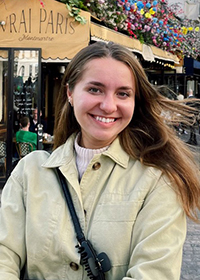Head-and-shoulders photo of a smiling young woman with long brown hair wearing a beige jacket and with a purse over her shoulder. She is standing outside Le Vrai Paris, a restaurant in Paris, France.
