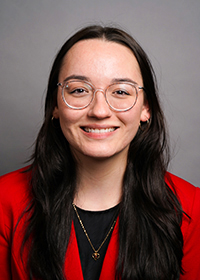 Head-and-shoulders photo of a smiling young woman with long dark brown hair and glasses wearing a red blazer and black top, in front of a gray background.