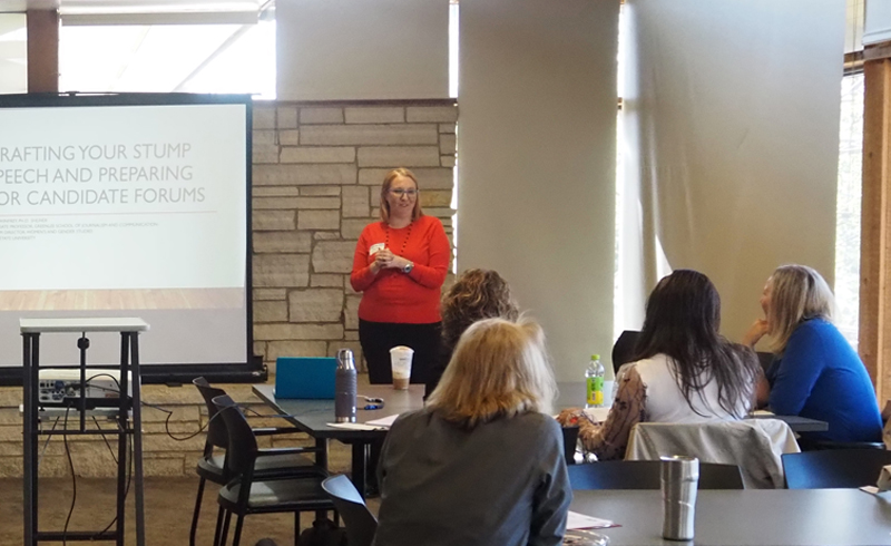 Women in orange sweater standing beside a screen at the front of a meeting room, while women seated at tables listen to her presentation.