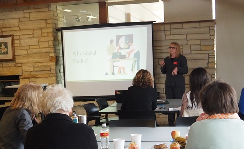 Woman in business suit standing beside a screen at the front of a meeting room. Two tables of meeting attendees are visible in front of her.