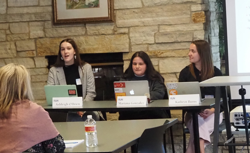 Three young women sitting at a table in the front of a meeting room, with a laptop open in front of each of them. The panelist on the left is speaking to the audience while the other two listen.