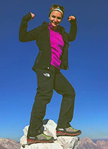 Young woman in hiking boots, black athletic pants and jacket, and magenta top standing on a rock formation and holding her arms in the air. The sky behind her is a deep blue and the rocky, mountainous landscape is visible in the distance.