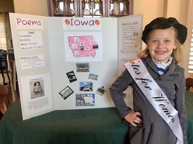 Young girl wearing a gray coat over a white blouse with a ruffled collar and blue bow, a black bonnet and a white sash that says "Votes for Women" poses beside a tri-fold form board with photos and information about Carrie Chapman Catt and Iowa.