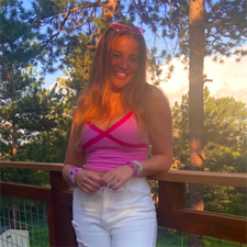 Young women with long brown hair wearing a pink top and white jeans, standing by a wooden deck rail with trees behind her.