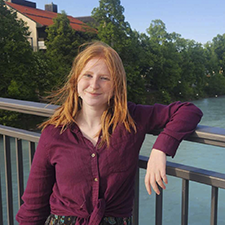 Young woman with shoulder-length red hair wearing a purple blouse, standing on a bridge over water, with trees in the background.