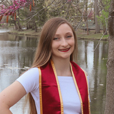 Young woman with long light brown hair standing beside Lake Laverne on Iowa State's campus, wearing a cardinal and gold graduation sash.