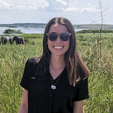 Young women with long brown hair wearing dark short-sleeved blouse and dark sunglasses, standing in a grassland with a lake and grazing animals behind her.
