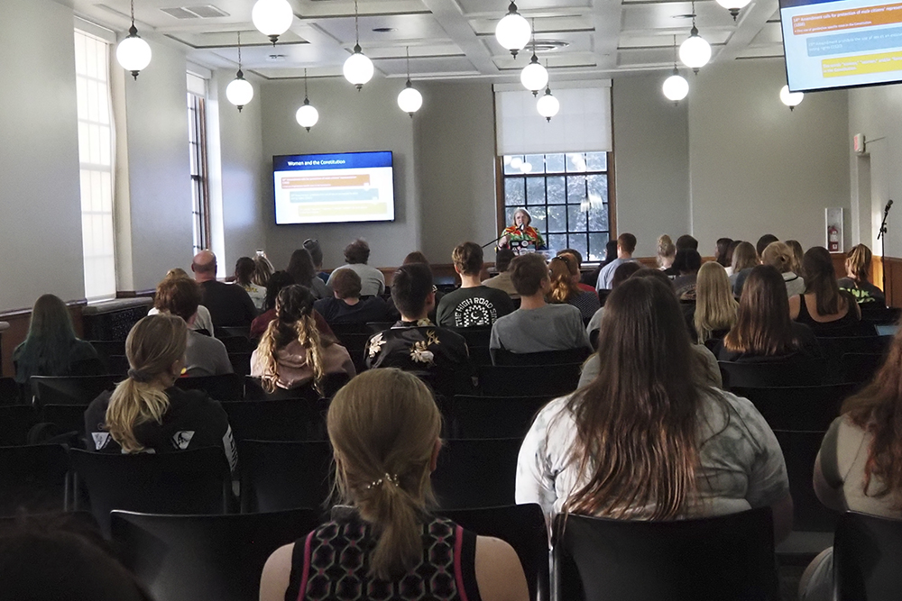 Photo taken from partway back in the room showing students listening to the speaker at the podium. A slide from the presentation is shown on the screen beside the speaker.