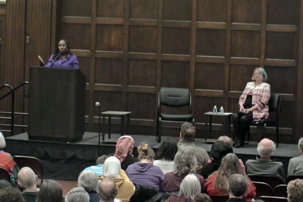 Sherrilyn Ifill stands at the podium in the Great Hall of the Memorial Union, while Karen Kedrowski is seated on the stage beside the chair that Ifill will take for the discussion.